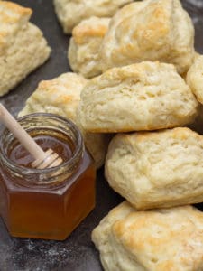 Homemade buttermilk biscuits piled high beside an open jar of honey with a honey wand