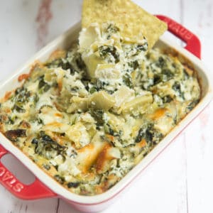 spinach artichoke dip with a tortilla chip in a red Staub baking dish on a white backdrop